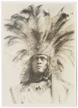 Head and shoulders of another Indigenous man in headdress with spreading feather crown. He wears a scarf knotted around his neck and a beaded blouse trimmed with fringe., Item 0001