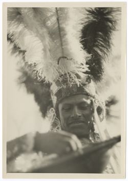 Head and shoulders of Indigenous man in headdress with beaded headband and pendants and upright feather crown. He is playing a guitar.