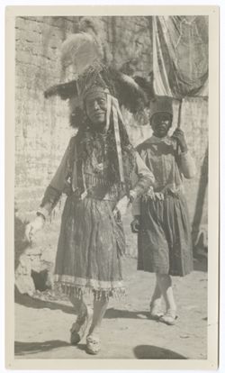 person in back carries banner on pole. On back, stamped in blue: "S.M. Eisenstein/Collective/Productions" and "Photo by/G. Alexandrov.", Two Indigenous people in procession, brick wall in background. Person in front wears bead and feather headdress